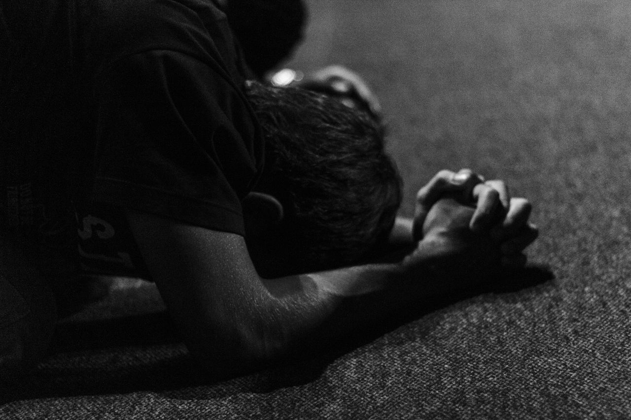 A black and white photograph capturing a person deeply engaged in prayer with clasped hands on the floor.