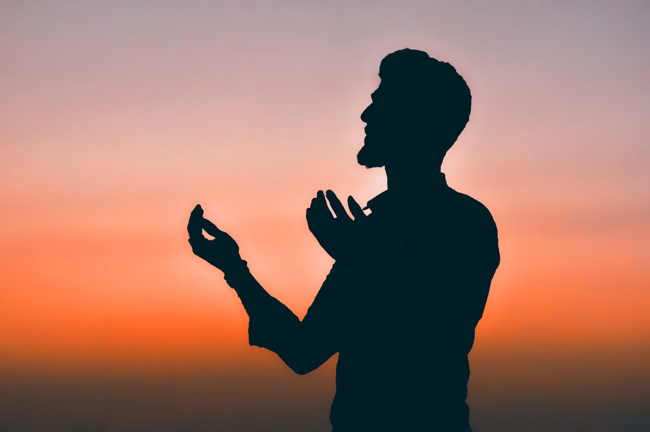 Silhouette of a man praying at sunset with hands raised against a vibrant sky.